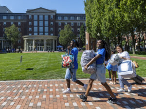Three smiling students carry bags, bedding, and a suitcase across a brick walkway on campus, with a residence hall and lawn visible in the background.