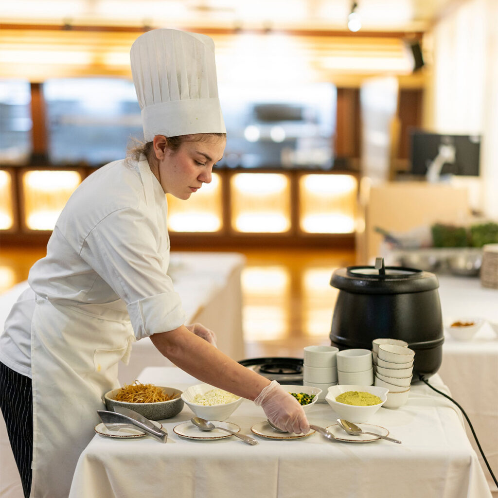 A culinary student prepares food on a table with various ingredients and small plates in a dining room.
