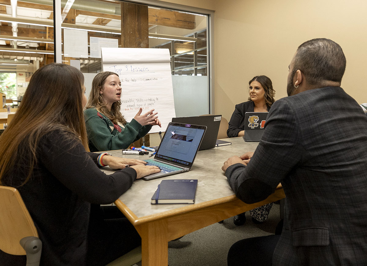A group of students engage in a collaborative meeting around a table, with one person speaking while others listen attentively.