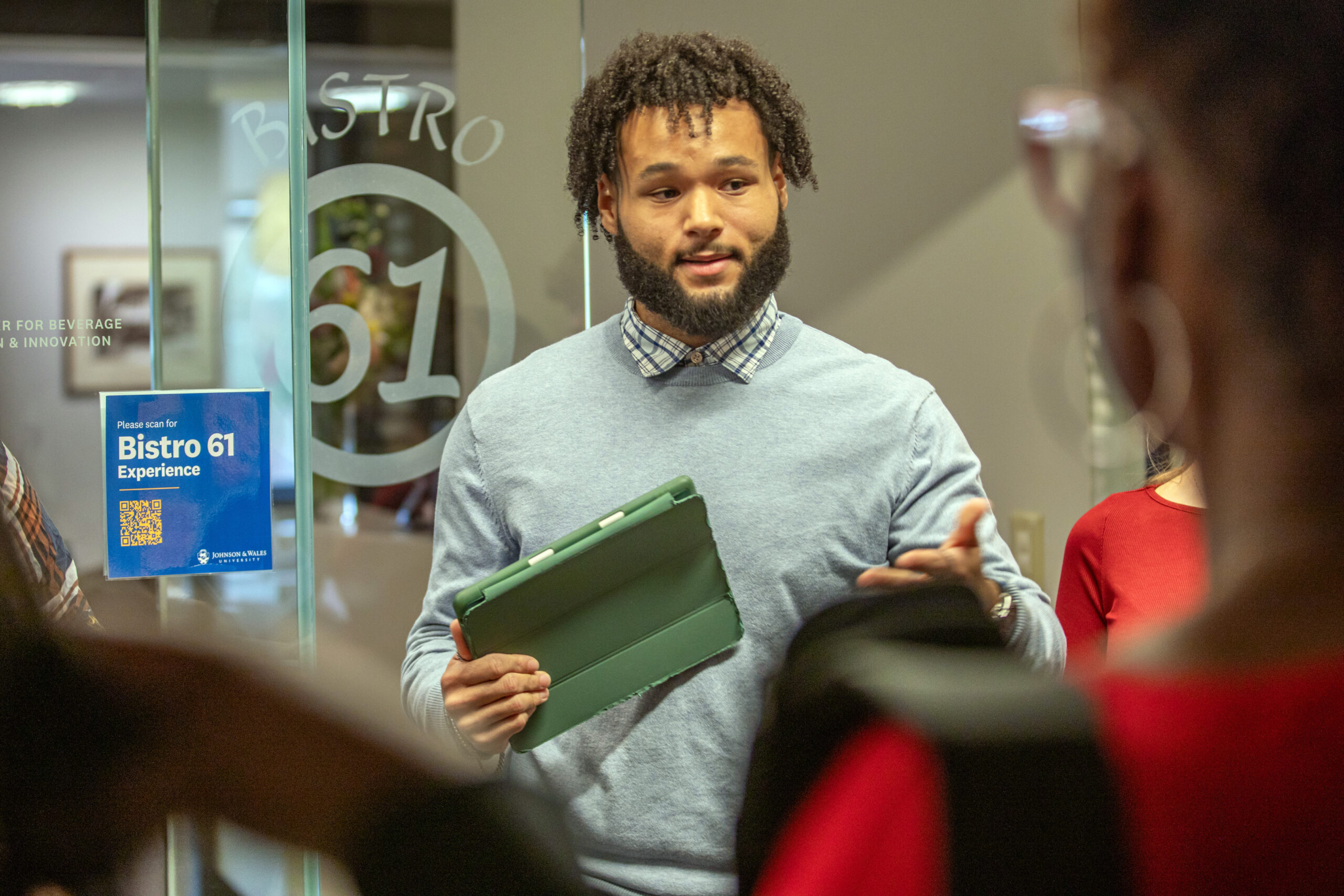 A person with curly hair and a beard, wearing a gray sweater over a collared shirt, holds a green tablet while speaking to others in a modern indoor setting near a glass door marked "Bistro 61."