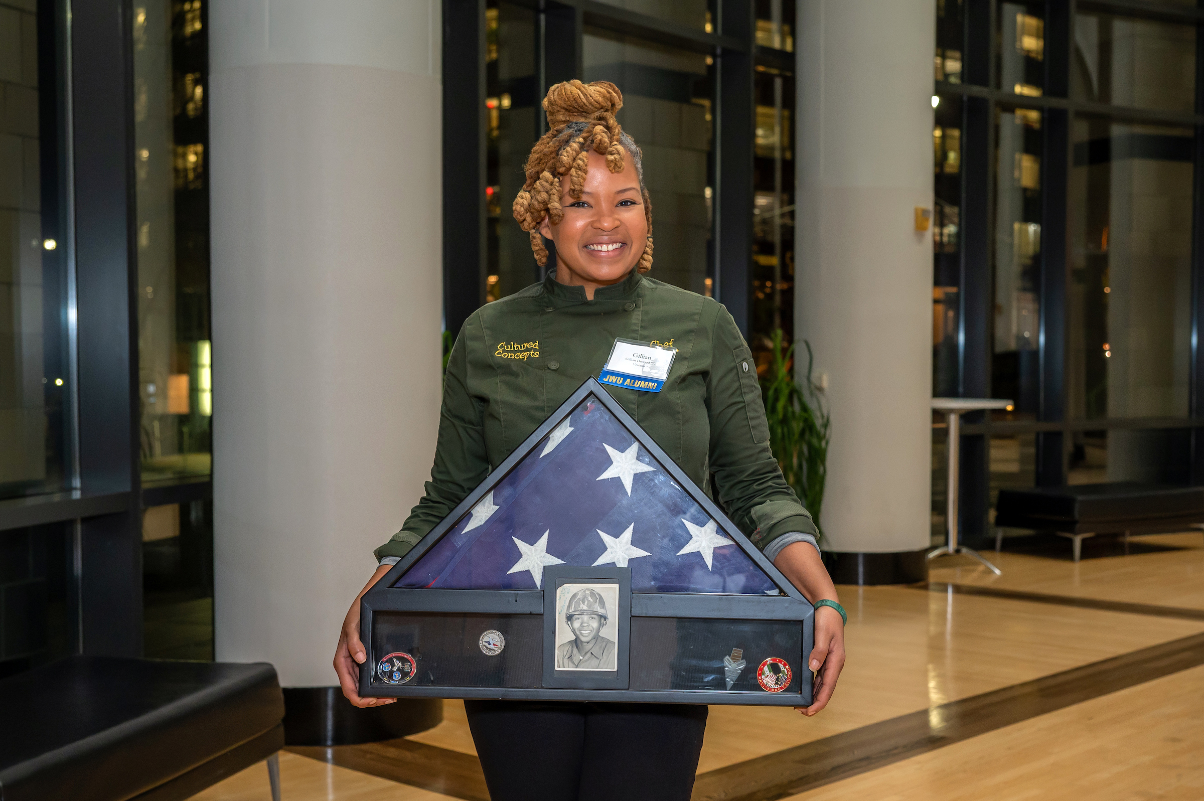 A person wearing a chef coat and JWU Alumni name badge stands indoors holding a framed American flag display case that includes medals and a black-and-white portrait.