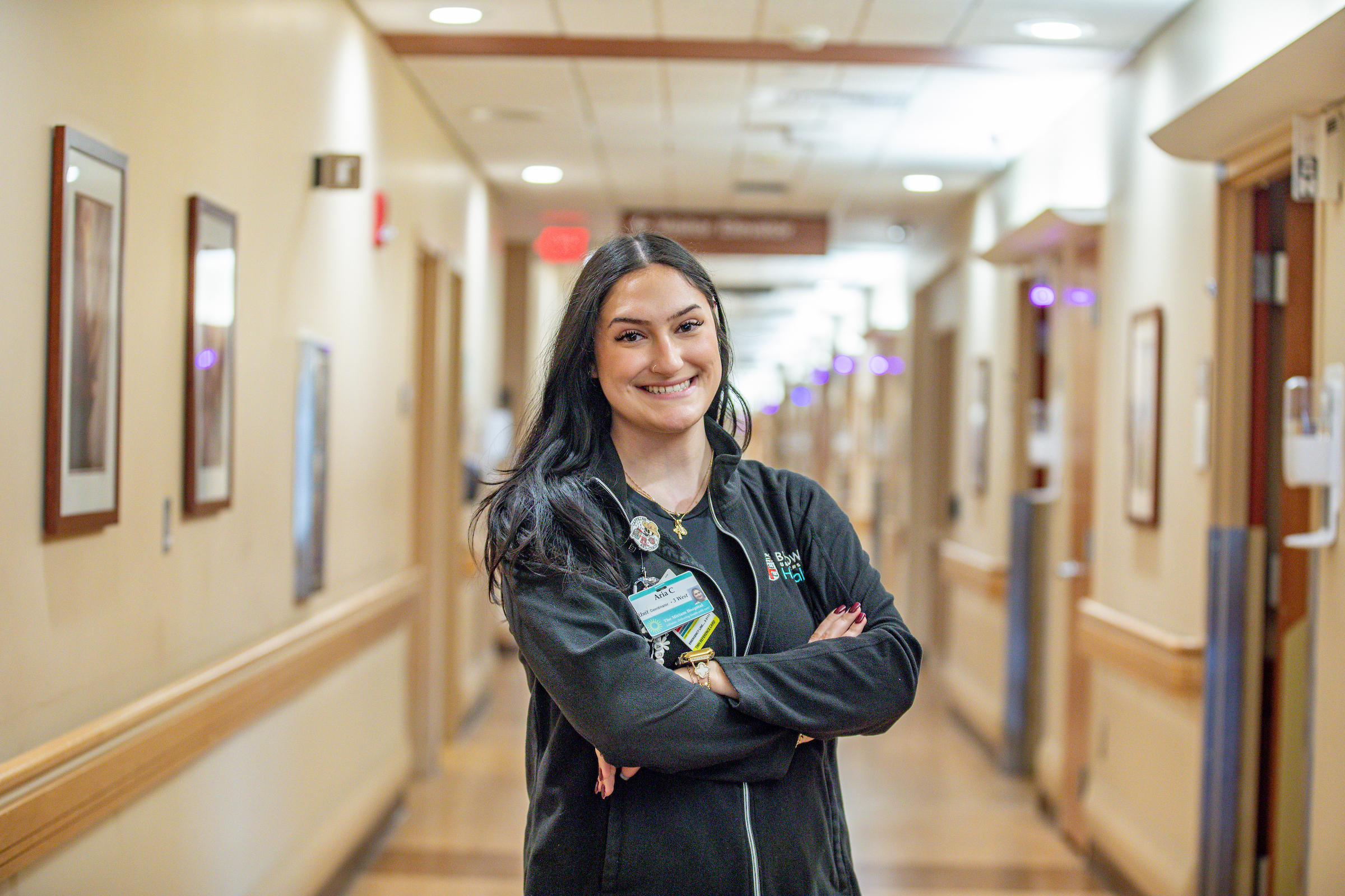 A healthcare worker stands with arms crossed in a hospital hallway, smiling at the camera.