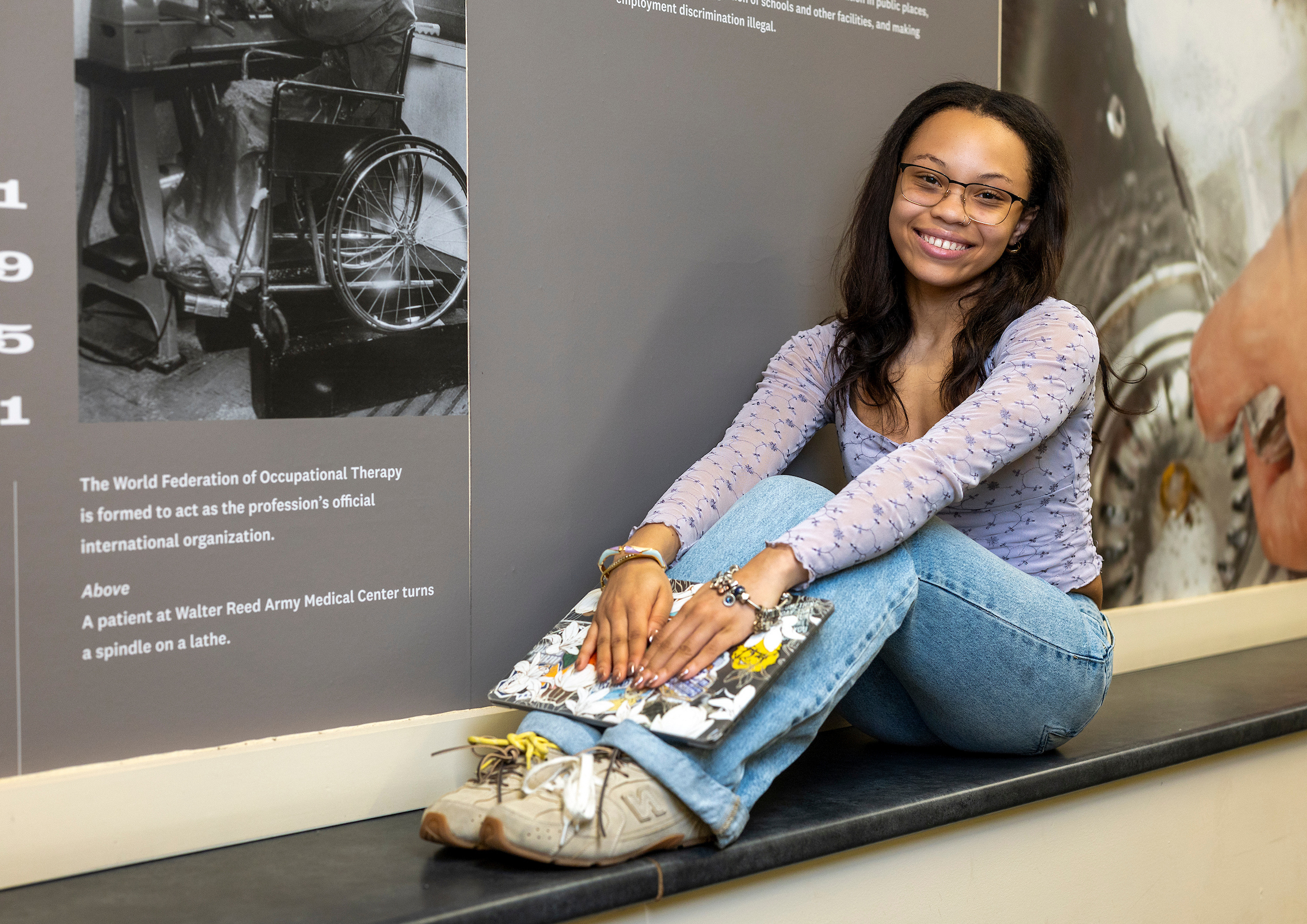 A young person with glasses sits on a bench, smiling, while holding a decorated notebook, in front of an informational exhibit wall.
