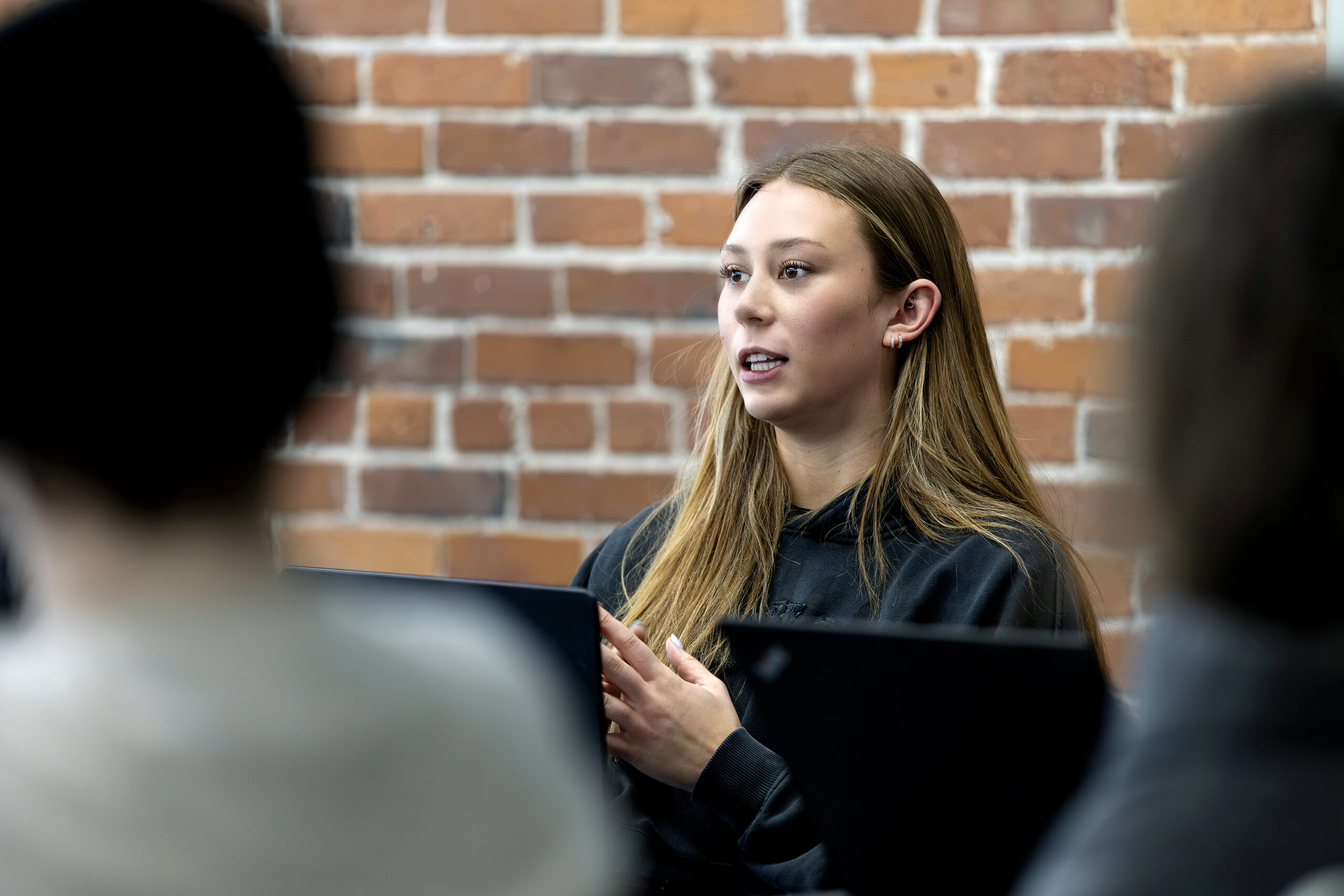 A person with long hair speaks to an audience while holding a tablet, standing in front of a brick wall.