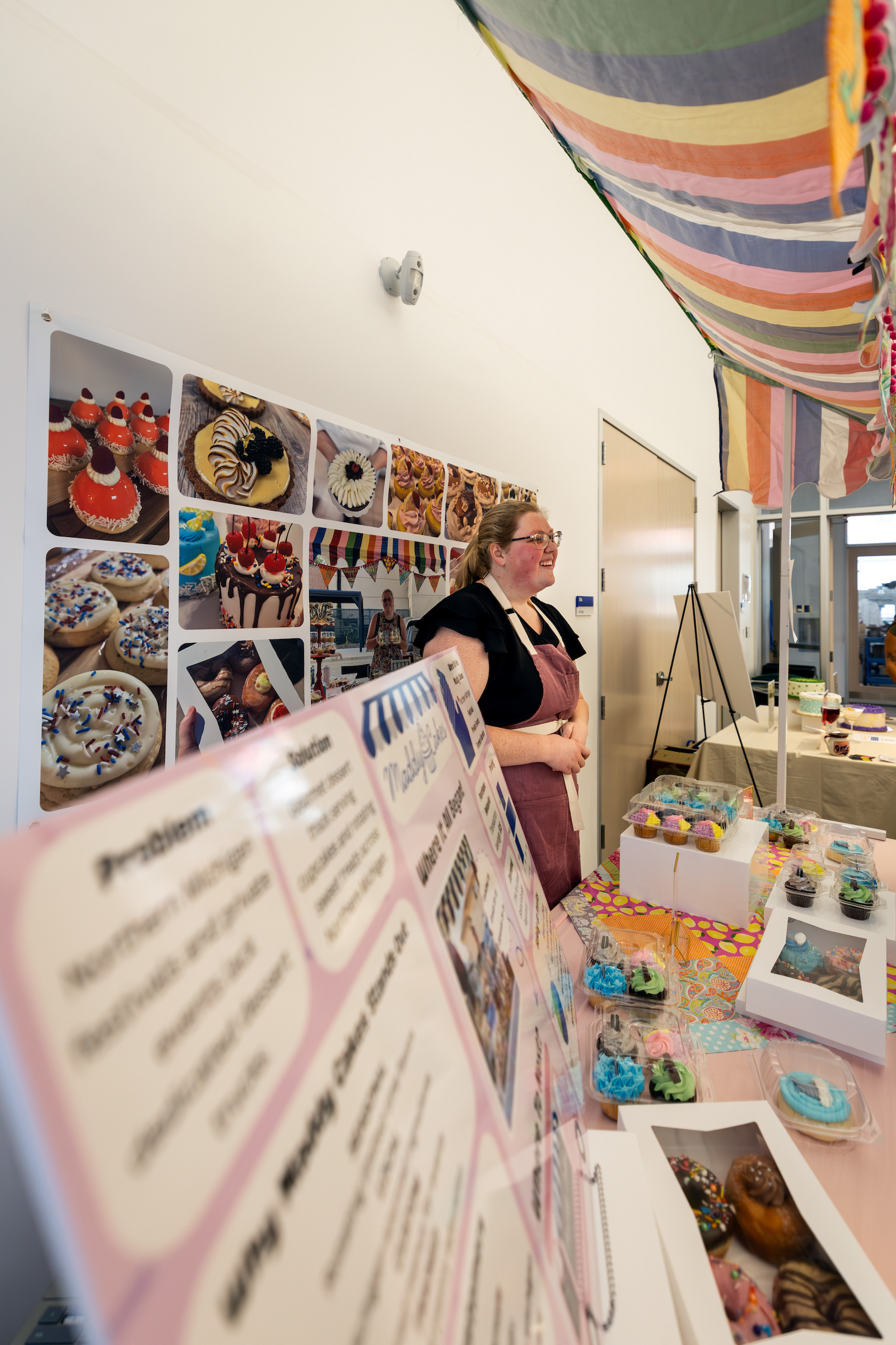A person in an apron stands beside a display of cupcakes and a presentation board at a colorful event booth.