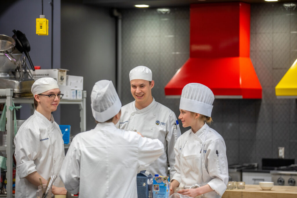 Four individuals in chef uniforms are engaged in conversation in a professional kitchen with a prominent red ventilation hood.