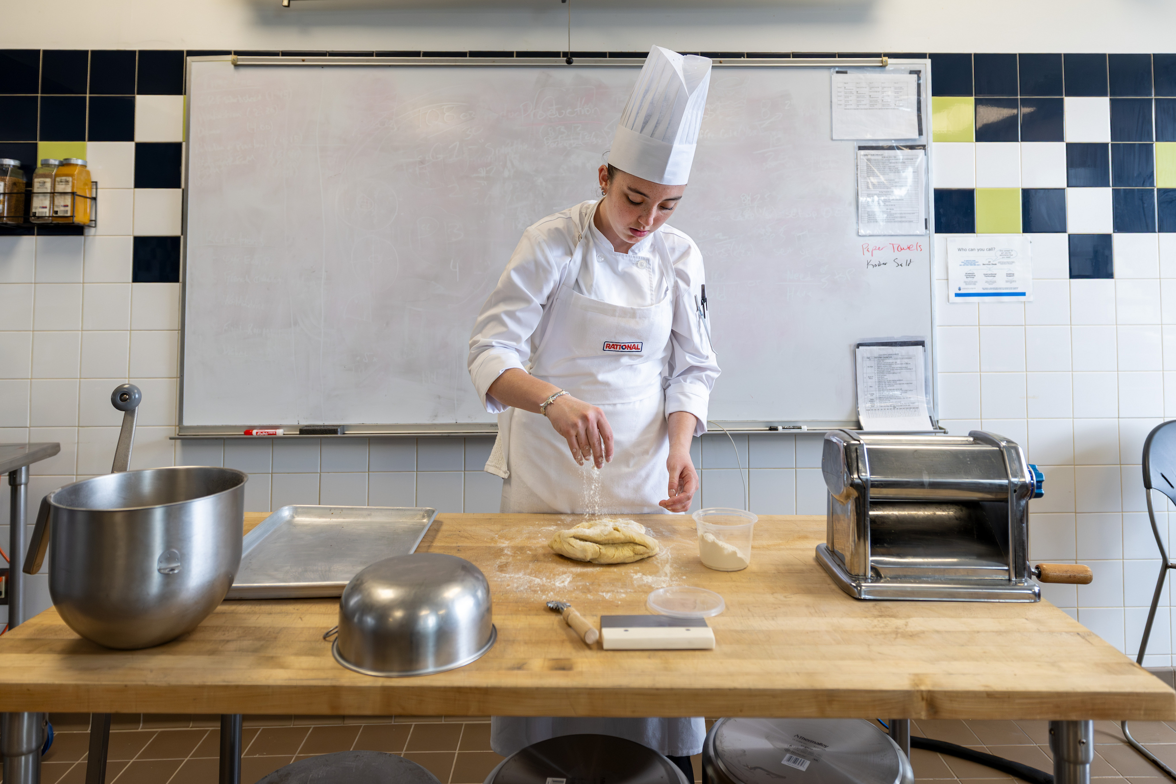 A culinary student in a chef's coat and toque sprinkles flour over dough on a wooden worktable.