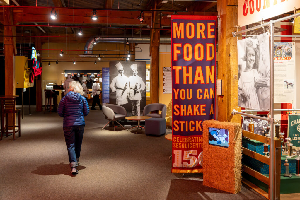 A visitor walks through a museum-style exhibit featuring historical photos, display cases with trophies, and a large banner reading “More Food Than You Can Shake a Stick,” under warm indoor lighting.