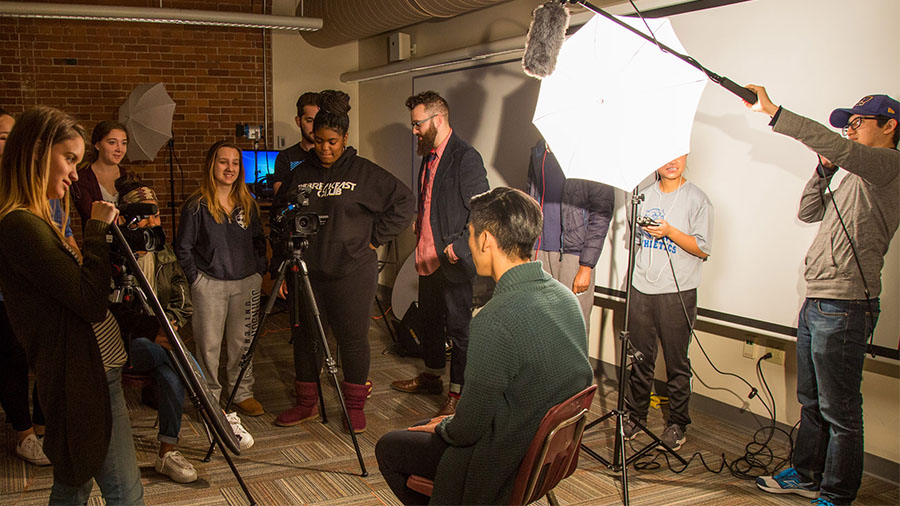 A group of students on a film set about to interview someone sitting in a chair.