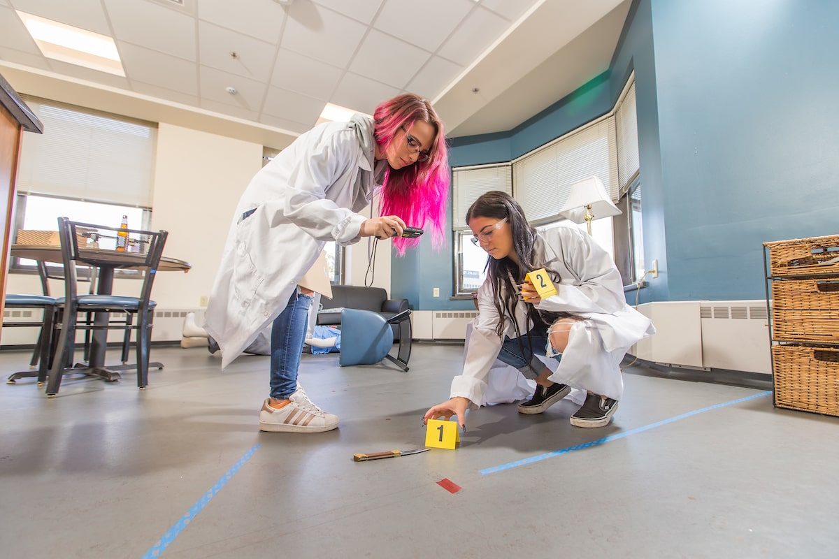 Two individuals in lab coats examine evidence markers on the floor of a room, one using a device and the other crouched near an item.