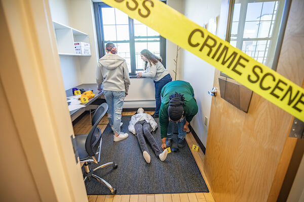 Students examine a mock crime scene with a body and evidence markers inside a room cordoned off by yellow tape.