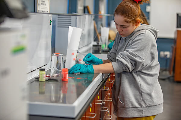 A person in a lab coat and gloves carefully handles a small container while working at a laboratory bench.