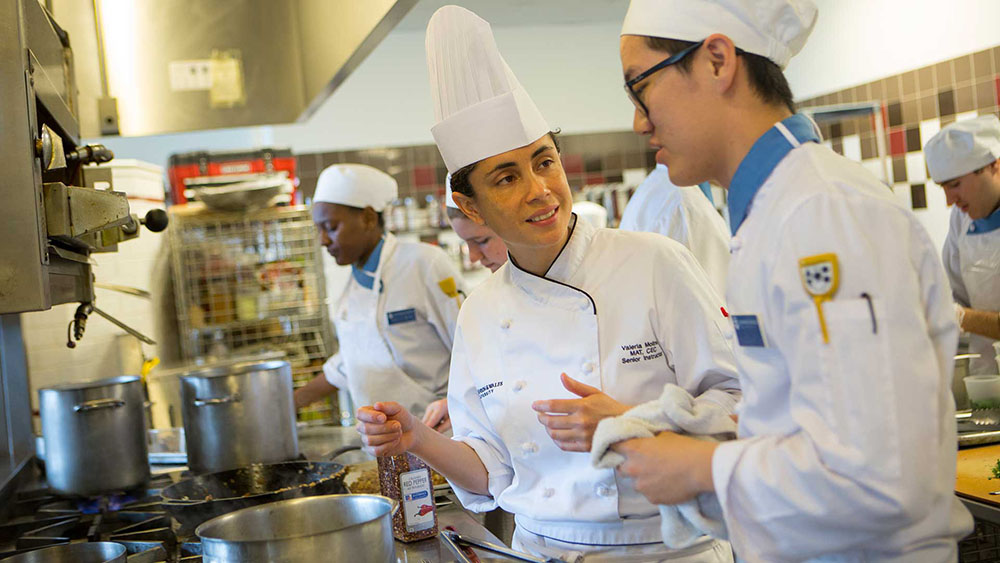 A culinary professor talking with a student in front of a stovetop.