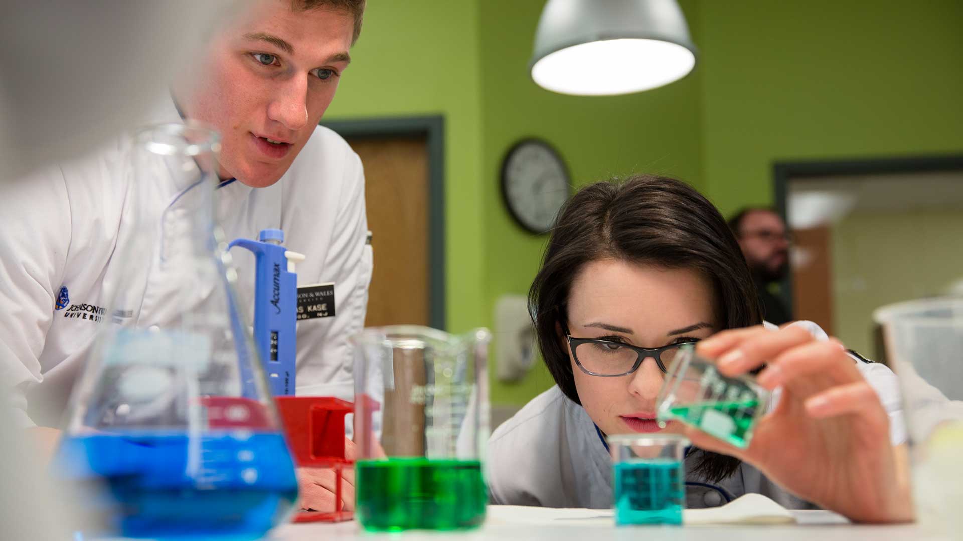 Student in white lab coat pours green liquid in a beaker in lab setting
