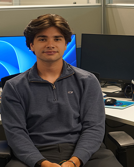 A student sits in a computer lab in front of multiple computer monitors, wearing a dark gray quarter-zip pullover and looking at the camera.