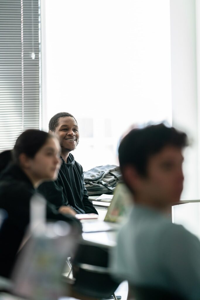 Three students in a classroom, with one in focus at the back-center, smiling toward the front of the room
