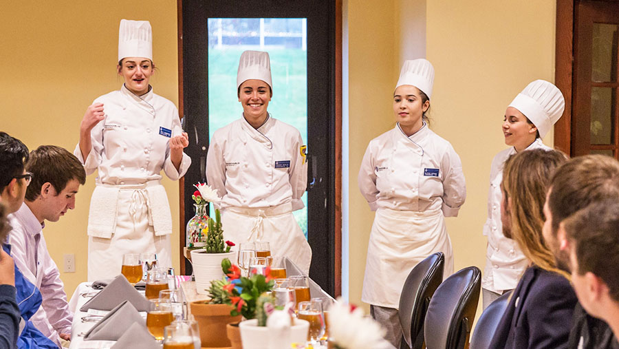 Four Culinary students present their dishes to a table of of guests in a dining room.