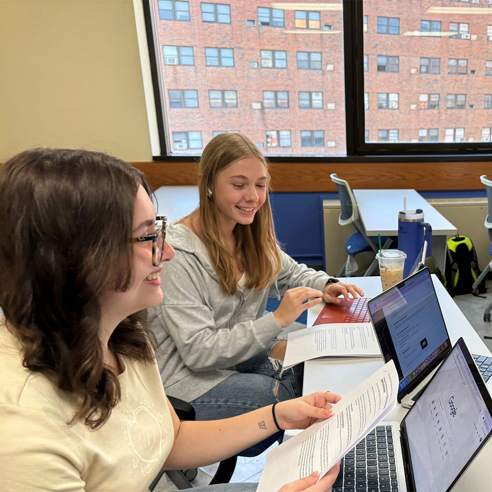 Two students smiling as they work on their laptops at a desk.
