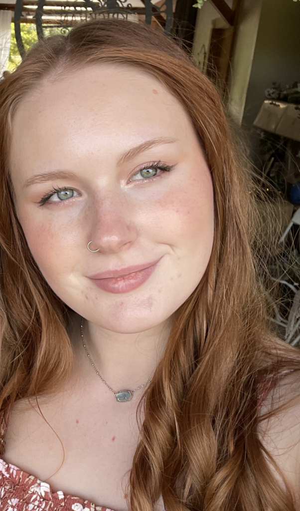 A young woman with long red hair and blue eyes, wearing a nose ring and a necklace, smiles at the camera in a well-lit indoor setting.