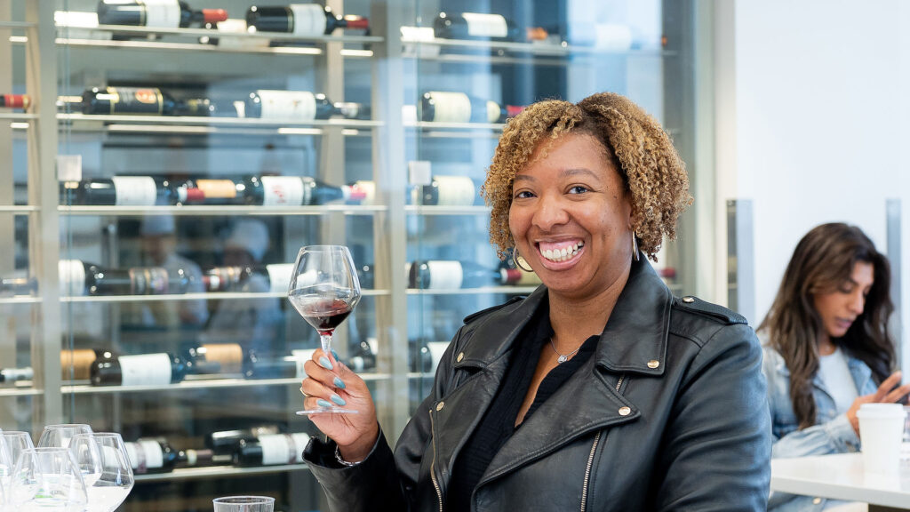 Smiling woman wearing holds a partially-filled red wine stem glass. Wine bottles are in a case behind her.