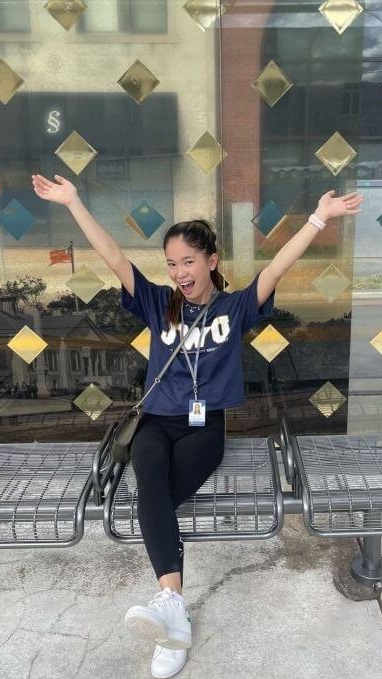 A smiling student in a JWU T-shirt sits on a bench outside a building with her arms raised, surrounded by decorative diamond-shaped window designs.