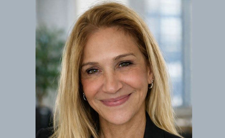 A smiling woman with long blonde hair and earrings poses for a headshot against a softly blurred indoor background.