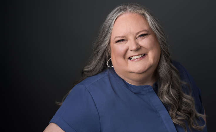 Smiling woman with long gray hair wearing a blue blouse stands against a dark background.