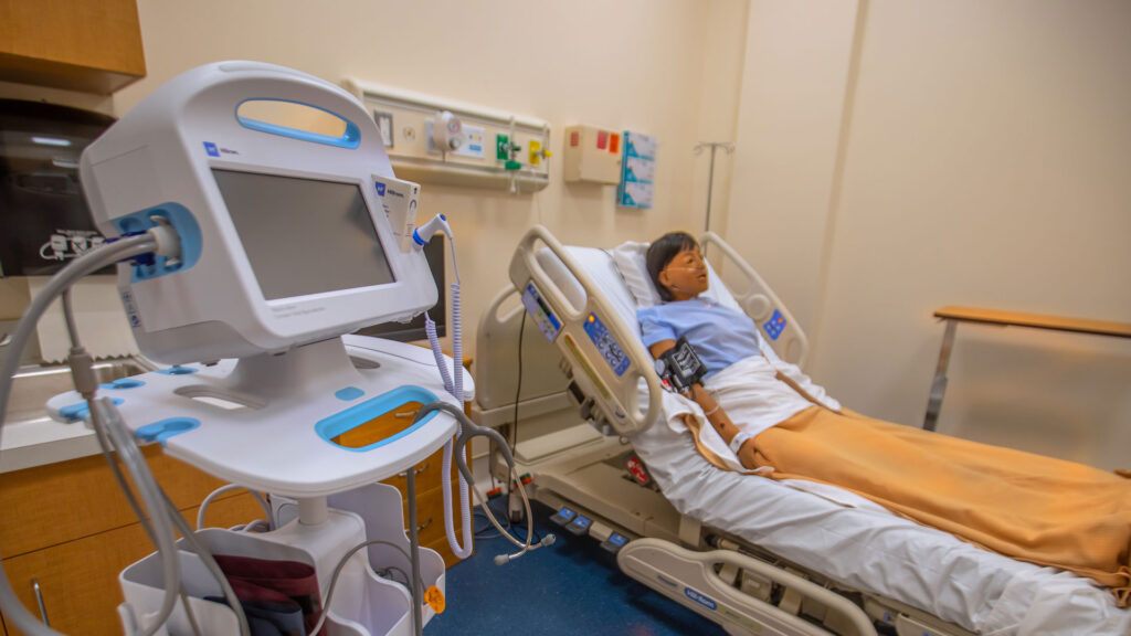 JWU nursing simulation center patient room with hospital bed, equipment, and dummy patient in bed