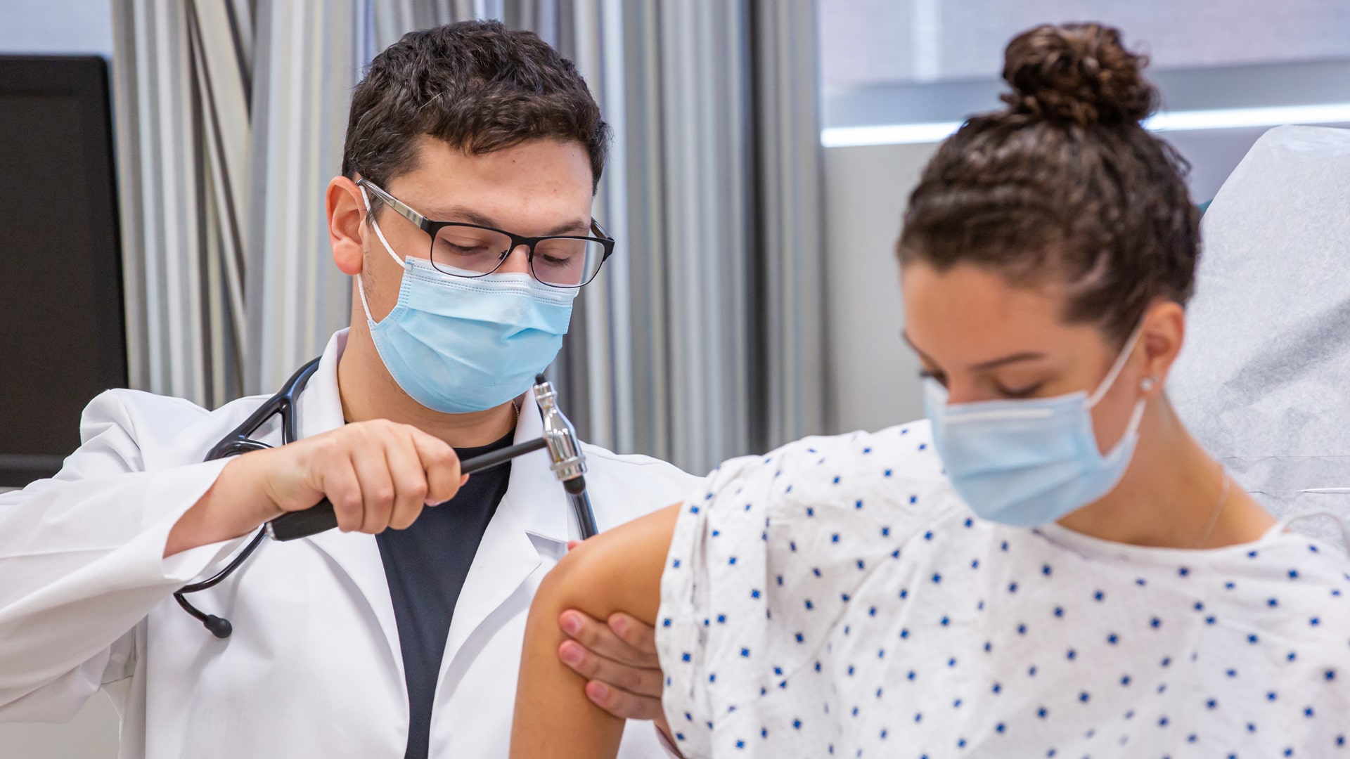 JWU nursing student in clinical student wearing facc mask and doctor's coat treating a patient in hospital gown