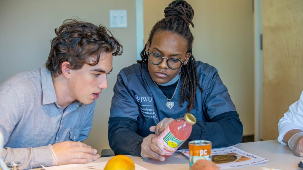 Two individuals collaborate at a table, examining a bottle of juice alongside food items and educational materials.