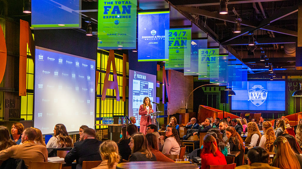 Woman on stage in front of screens, standing in front of large audience