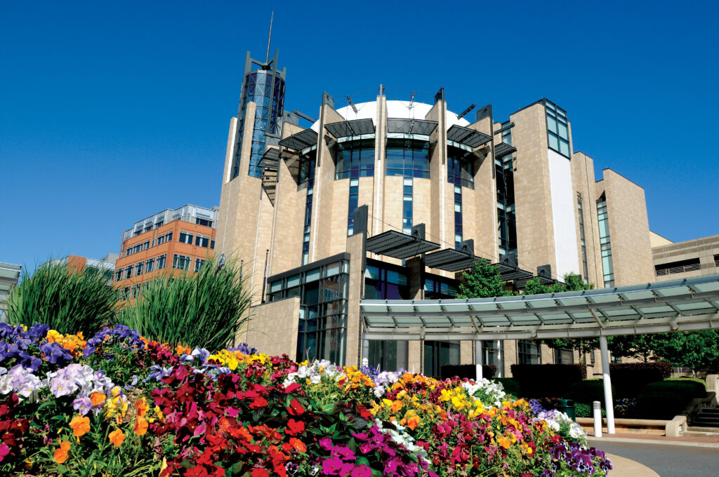 A modern building with a distinctive circular design and glass accents stands under a clear blue sky, surrounded by vibrant flower beds and greenery.