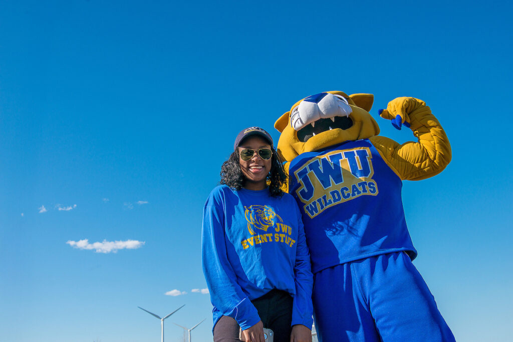 A student in a blue Johnson and Wales shirt stands beside Wildcat Willie under a clear blue sky.