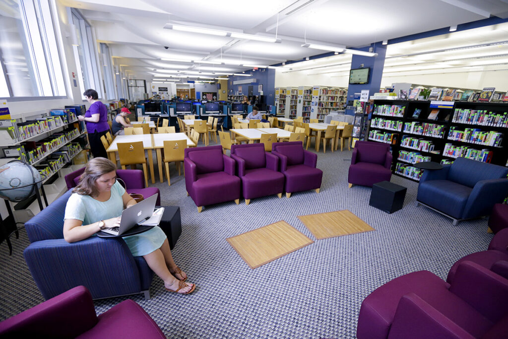 A person uses a laptop while seated in a comfortable chair in the Charlotte library featuring bookshelves, study tables, and plush seating.