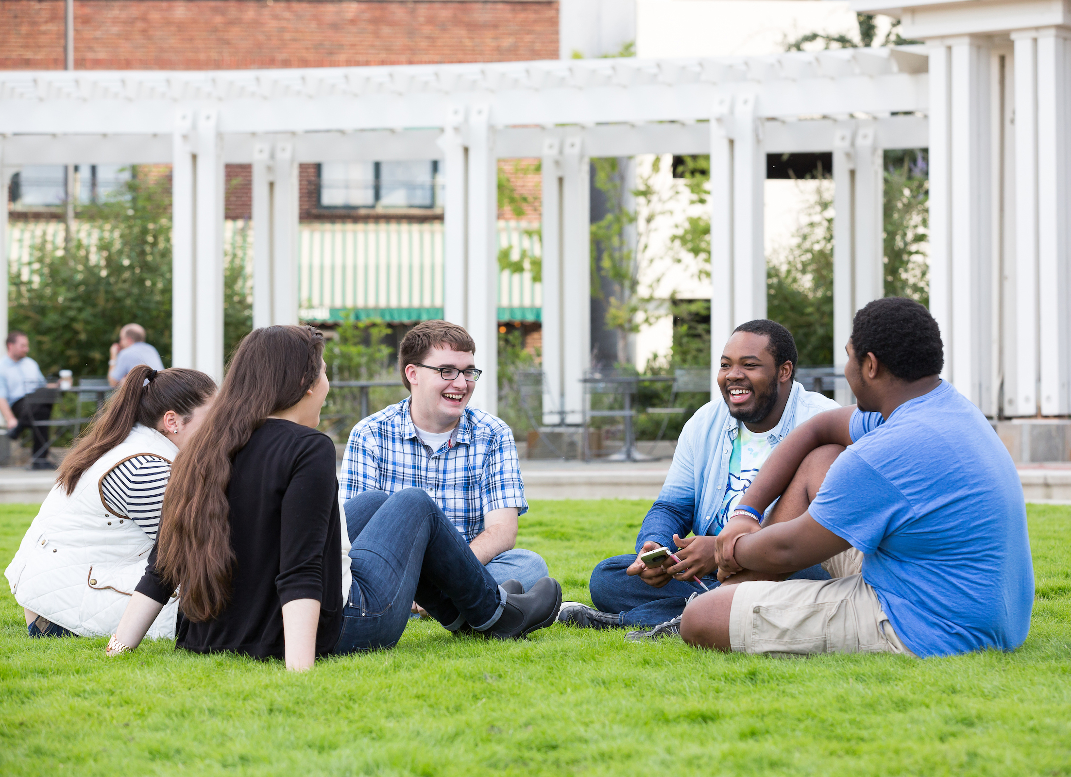 A group of young people are sitting on grass, laughing and chatting together in an outdoor setting.