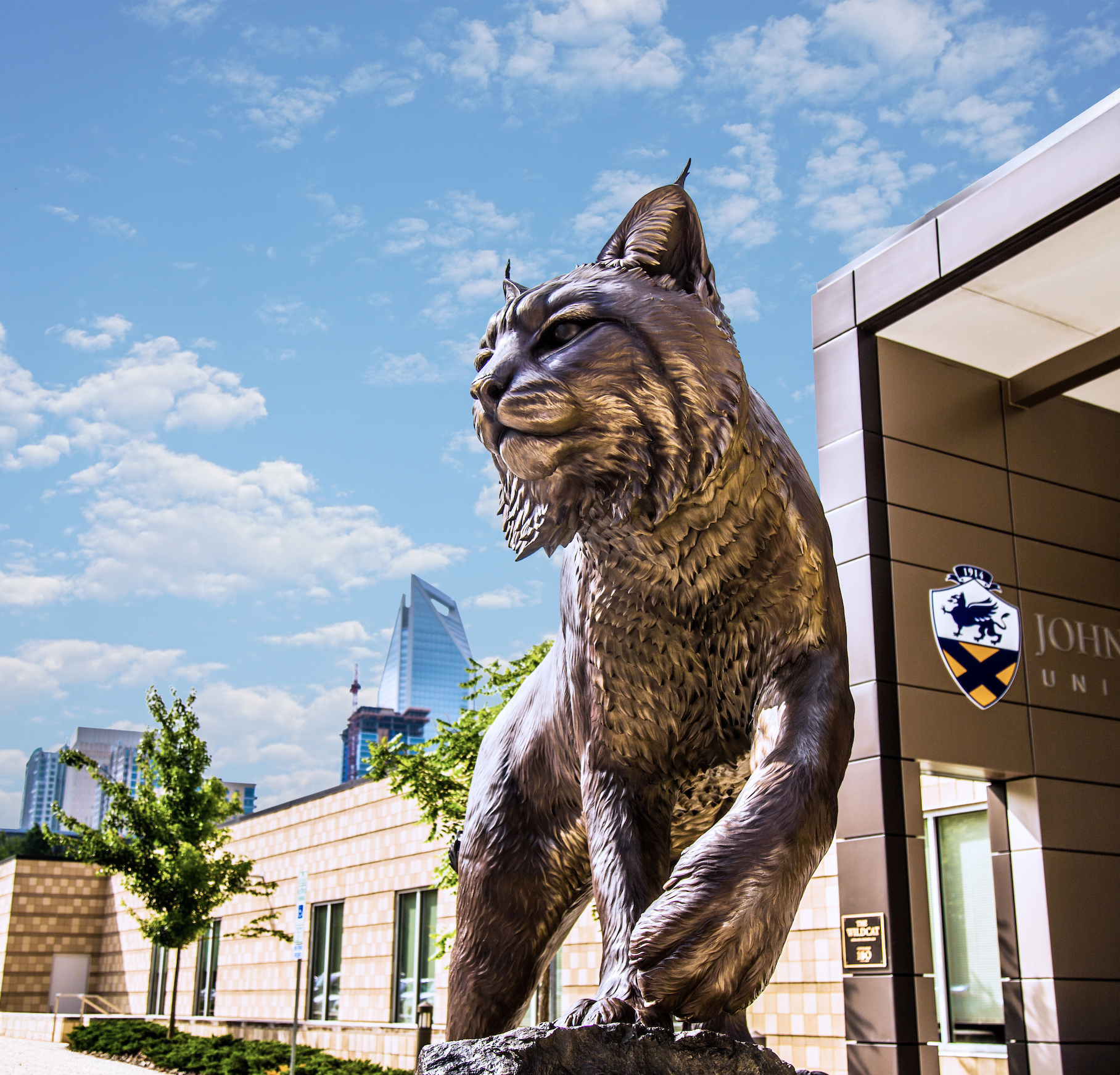 A bronze statue of a lynx stands prominently outside Johnson & Wales University, with modern buildings and a blue sky in the background.