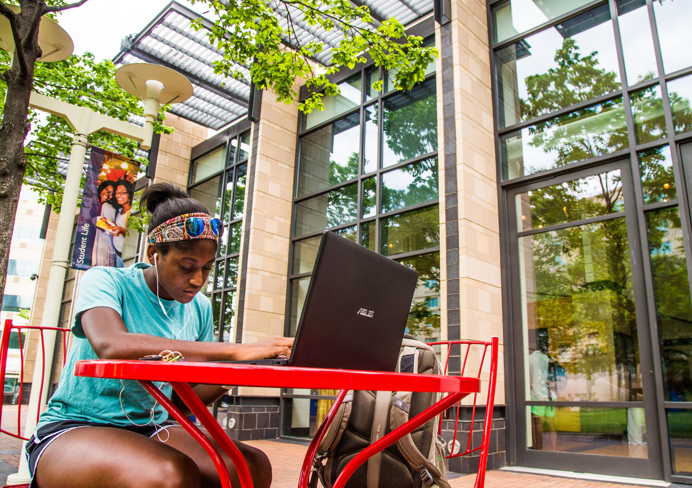 A person sits at a red outdoor table, using a laptop while wearing earphones and a headband, with a modern building and green trees in the background.