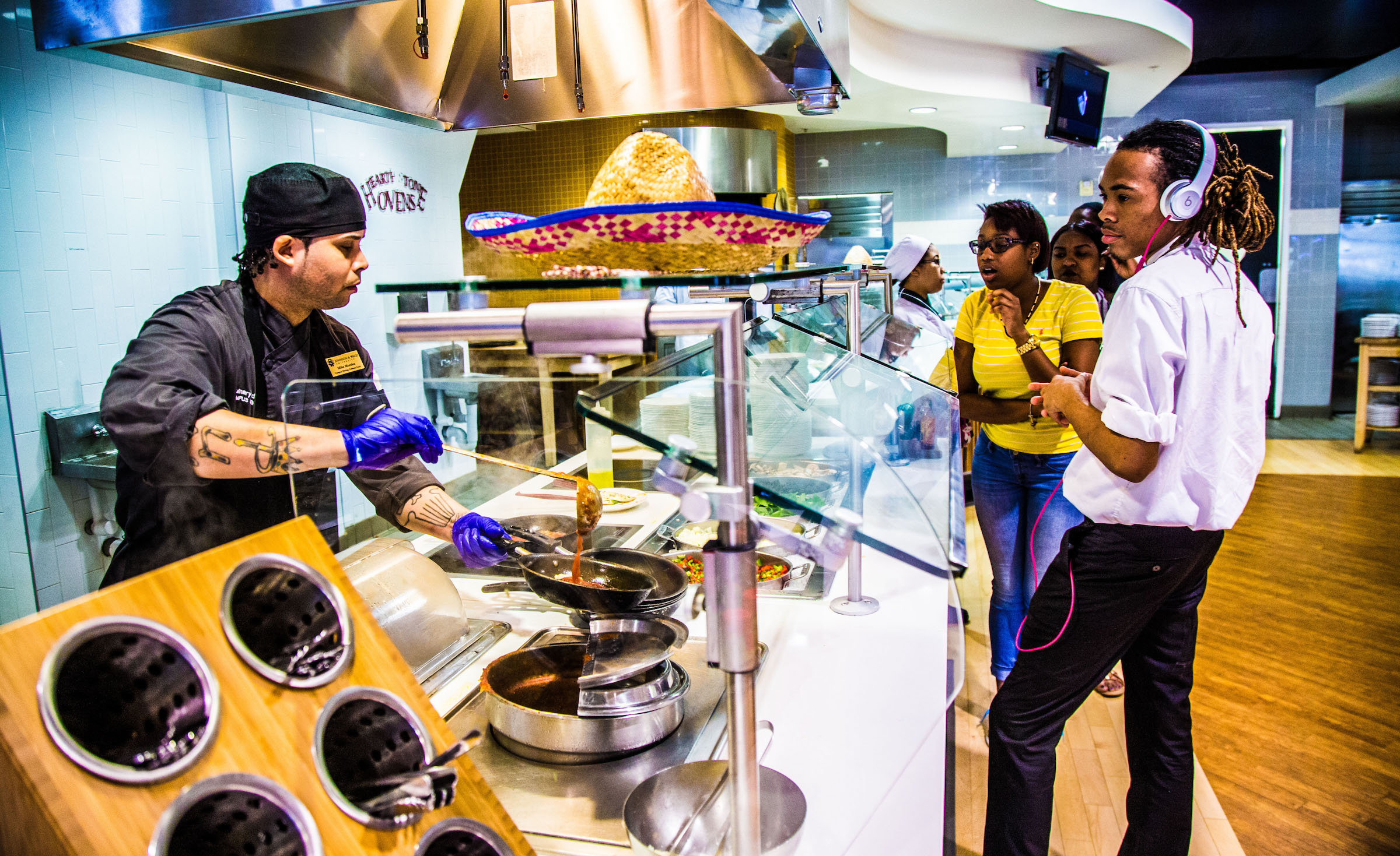 A chef prepares food at a counter while customers observe, one wearing headphones.