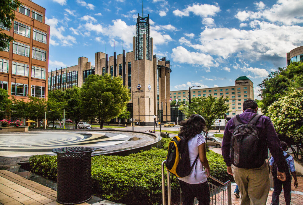 People walk through a sunny urban plaza with modern buildings, green trees, and a circular water feature under a bright blue sky.