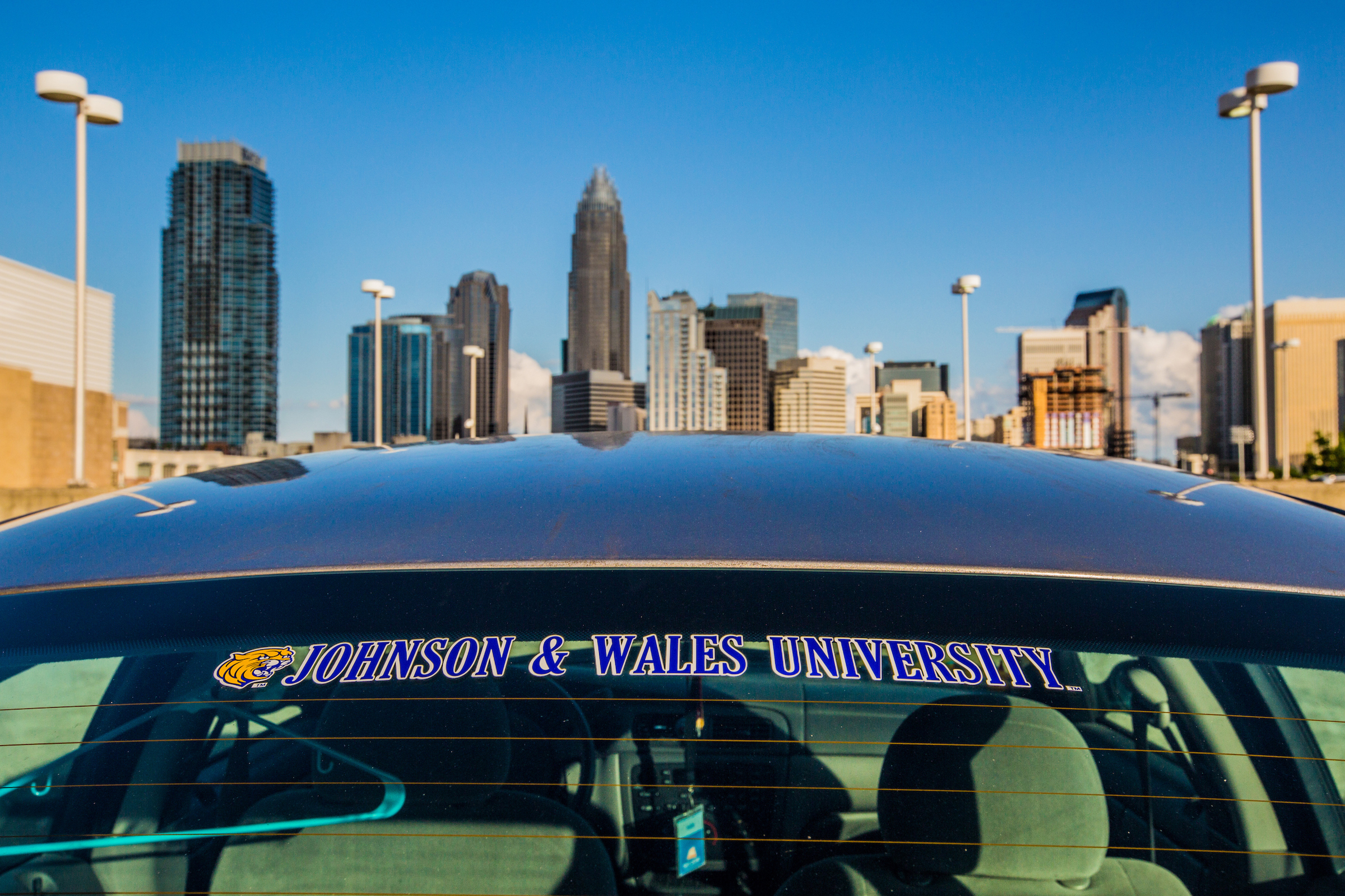 A car with a "Johnson & Wales University" decal is parked in front of a city skyline under a clear blue sky.
