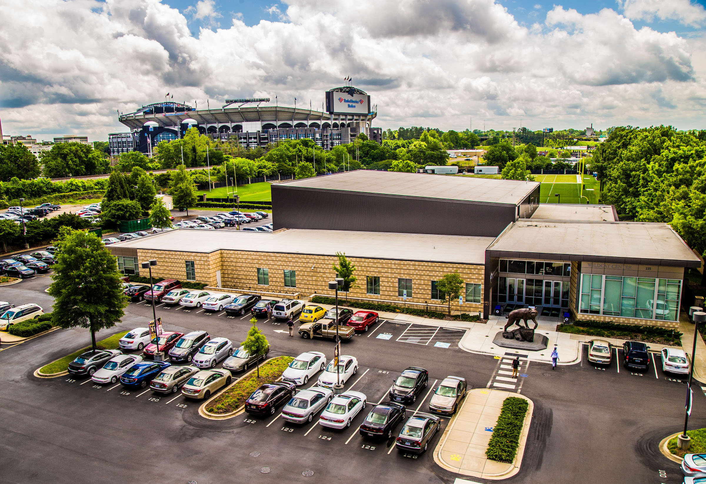 An aerial view of a parking lot with cars and a building in front of a large stadium under a cloudy sky.