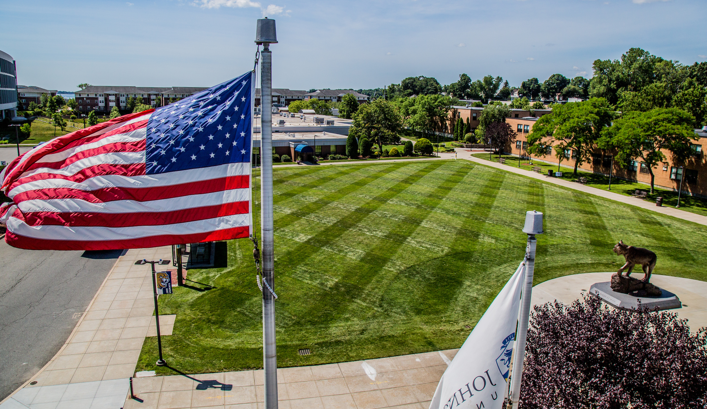 A large American flag waves in the breeze over a well-maintained green lawn, with campus buildings and a statue visible in the background.