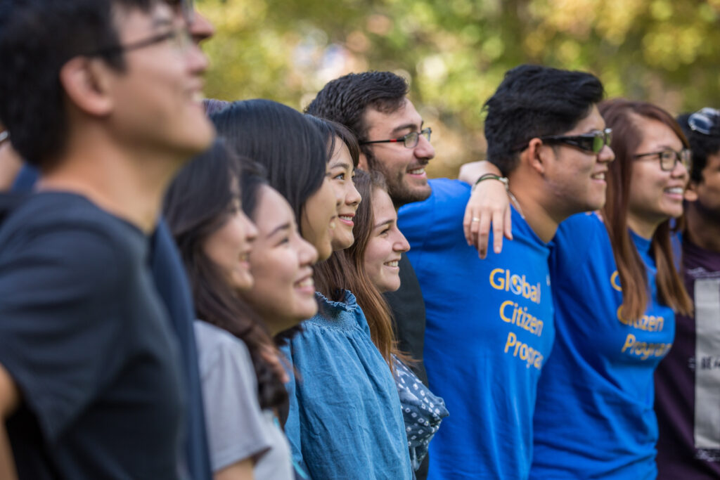 A diverse group of individuals, some wearing "Global Citizen Program" shirts, smile and stand together outdoors.