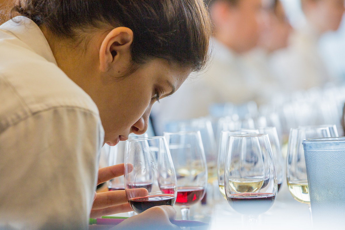 A person leans over a table filled with wine glasses, closely examining a glass of red wine.
