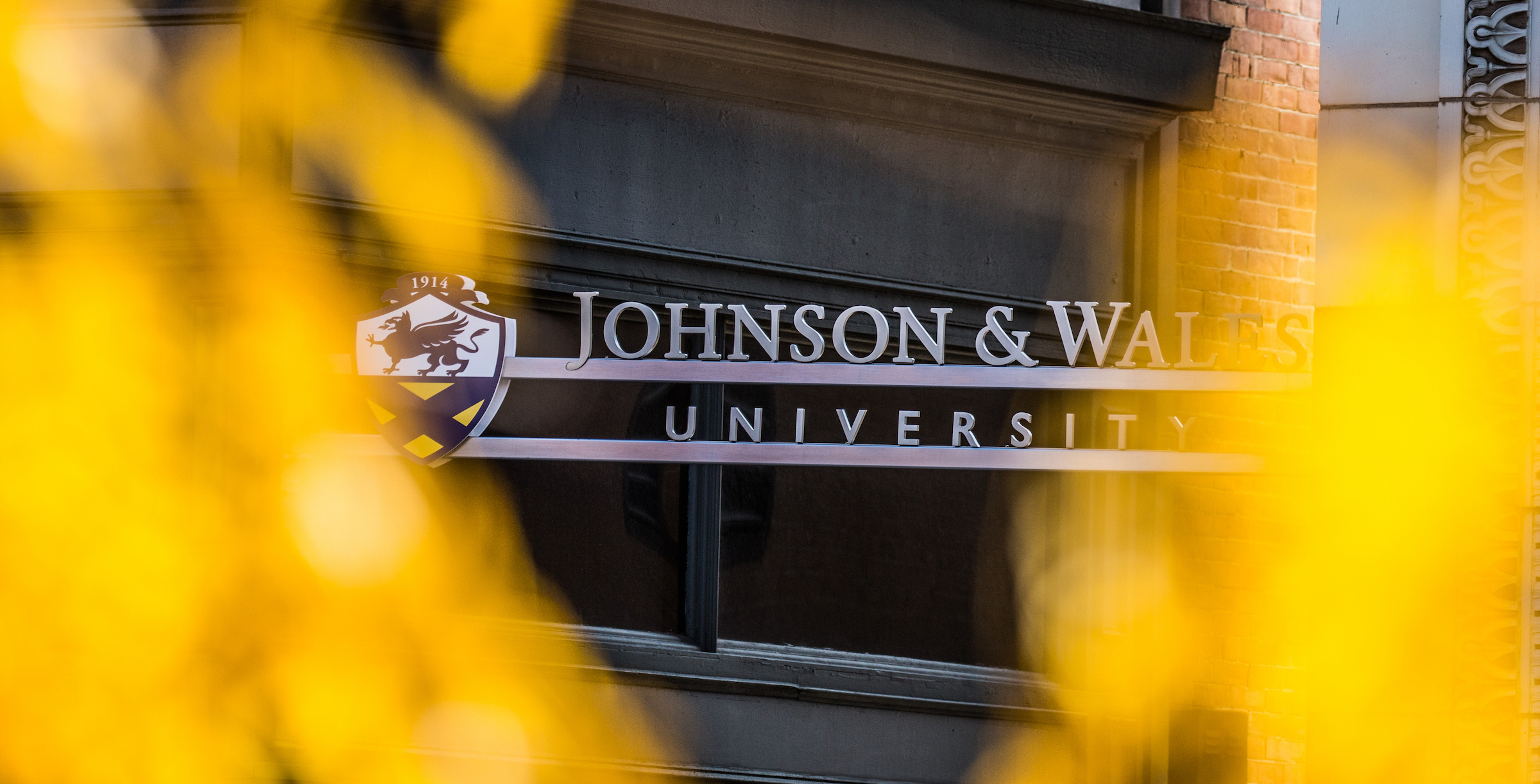 A sign reading Johnson and Wales University is framed by blurred yellow foliage in the foreground.