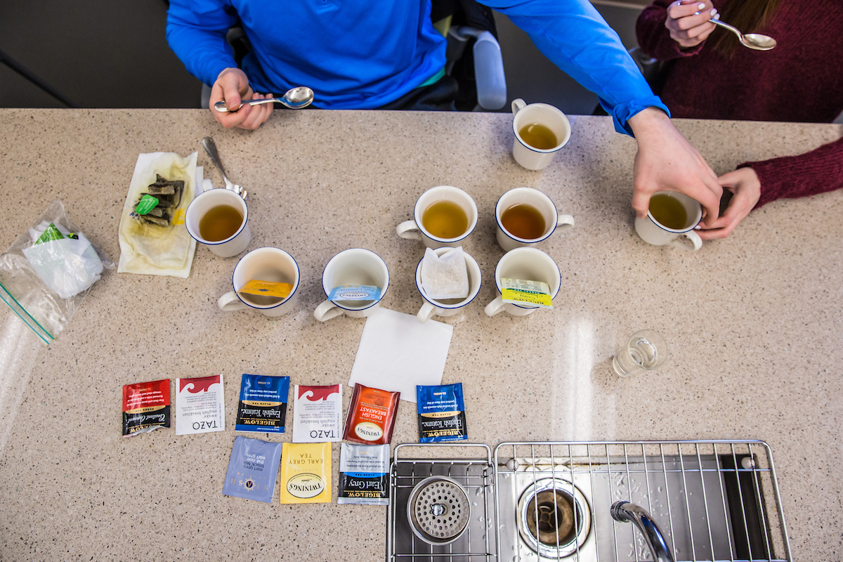 Two people prepare tea using various tea bags and spoons at a kitchen counter with multiple cups and a sink.