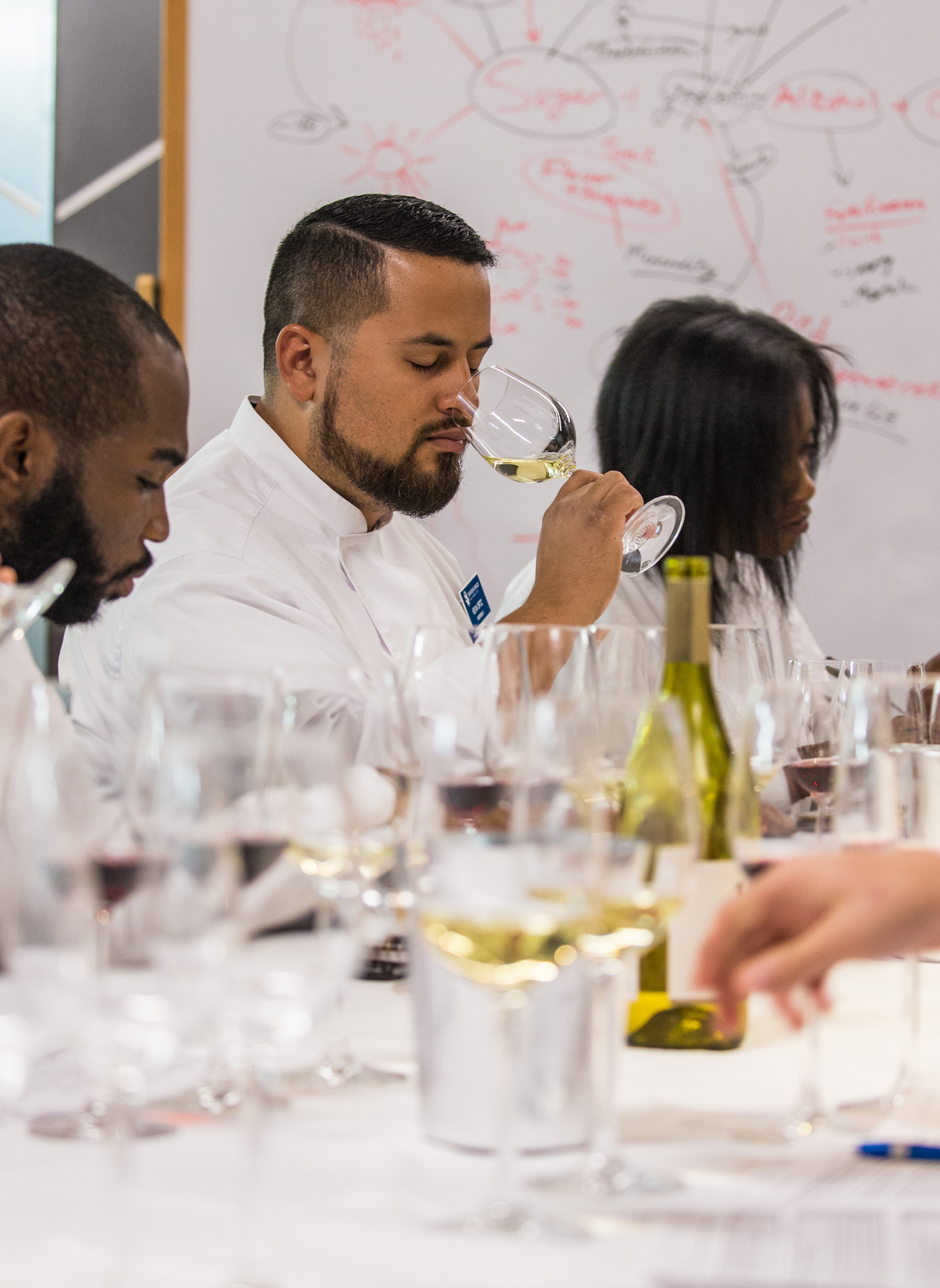 A person in a white chef’s coat smells wine from a glass during a tasting session, with others seated around a table filled with wine glasses and a whiteboard displaying wine tasting notes in the background.