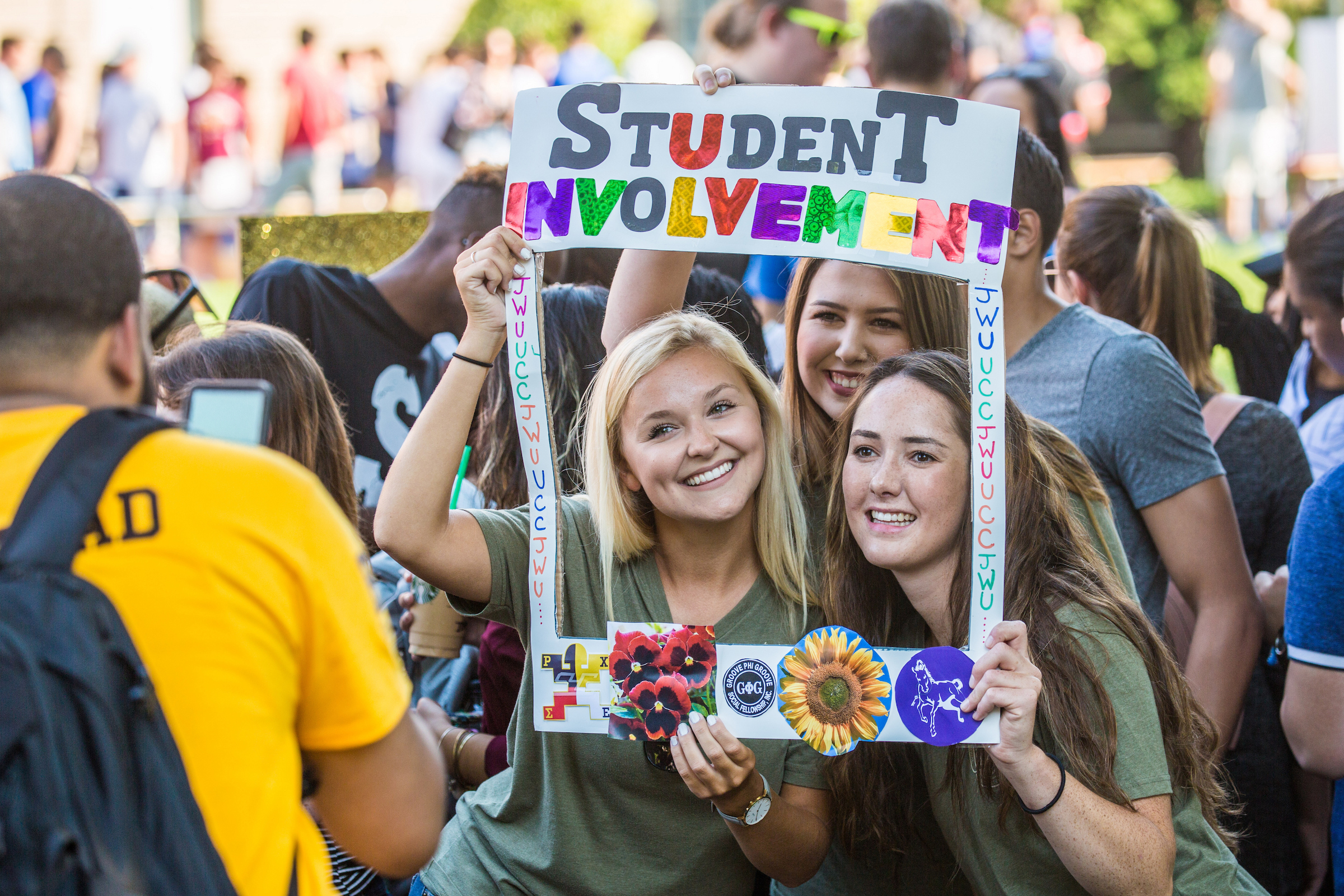 Three young people smile while holding a colorful sign that reads "STUDENT INVOLVEMENT" at an outdoor gathering.