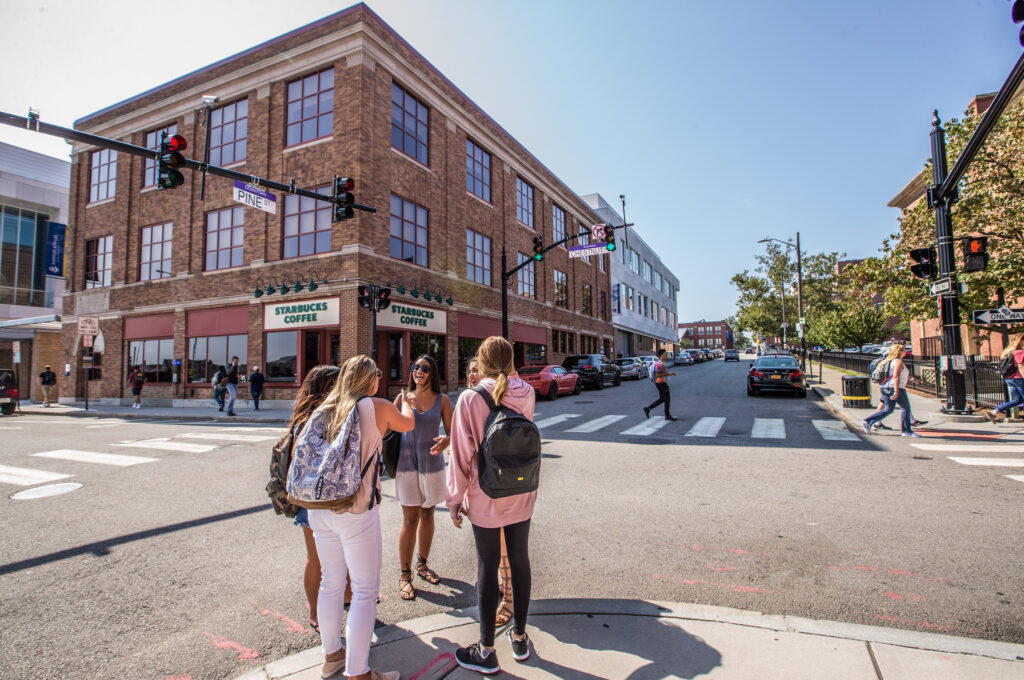 A group of people stands at a crosswalk near a brick building with a Starbucks Coffee sign on a sunny day.