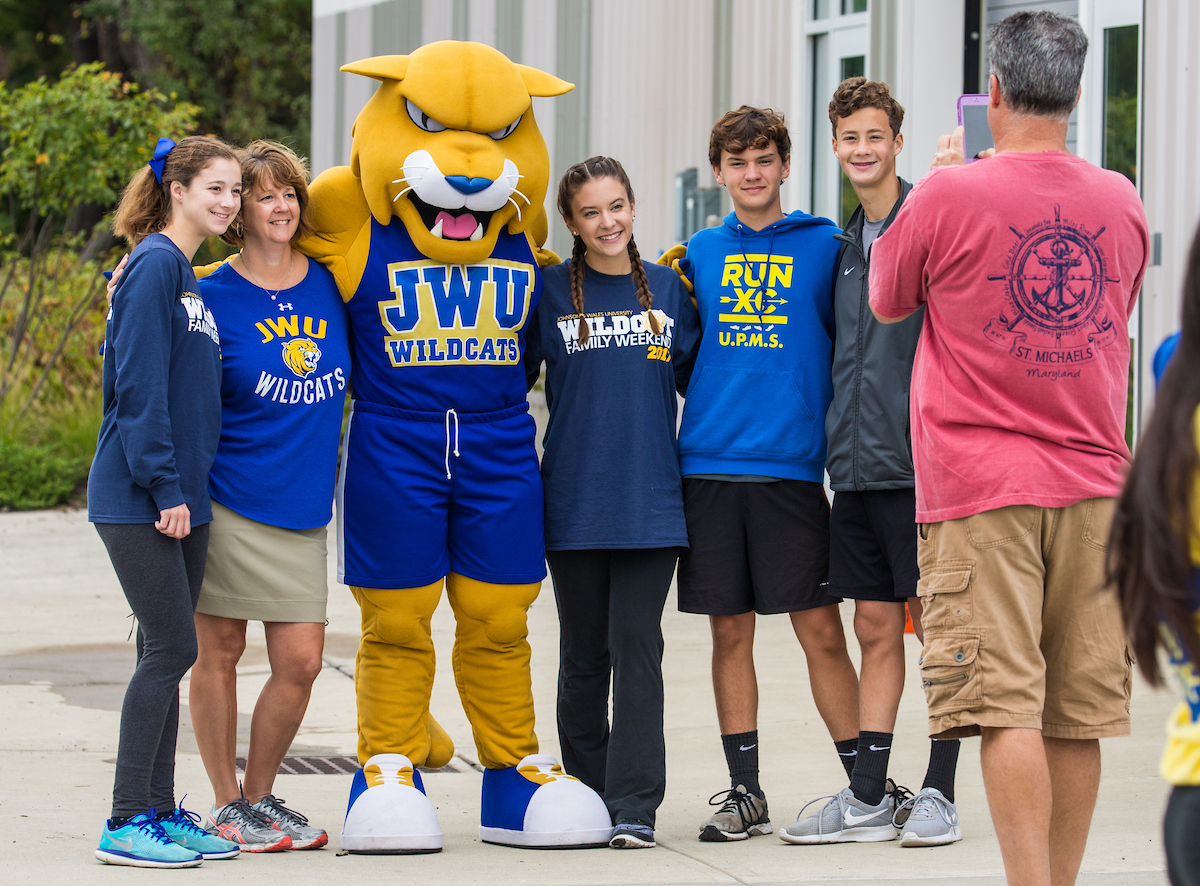 A group of people, including a large yellow mascot, pose together outdoors for a photo, with one person capturing the moment on their phone.